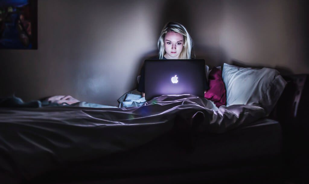 Young woman sitting on bed with laptop open in the dark
